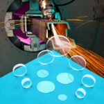 An array of clear spherical laser debris shield sit on a blue table. a photo of a laser cutting metal is in the background.