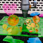 Various sizes of orange Zinc Selenide laser lenses stand on a piece of green acrylic. The acrylic is being cut into different shapes by a laser.