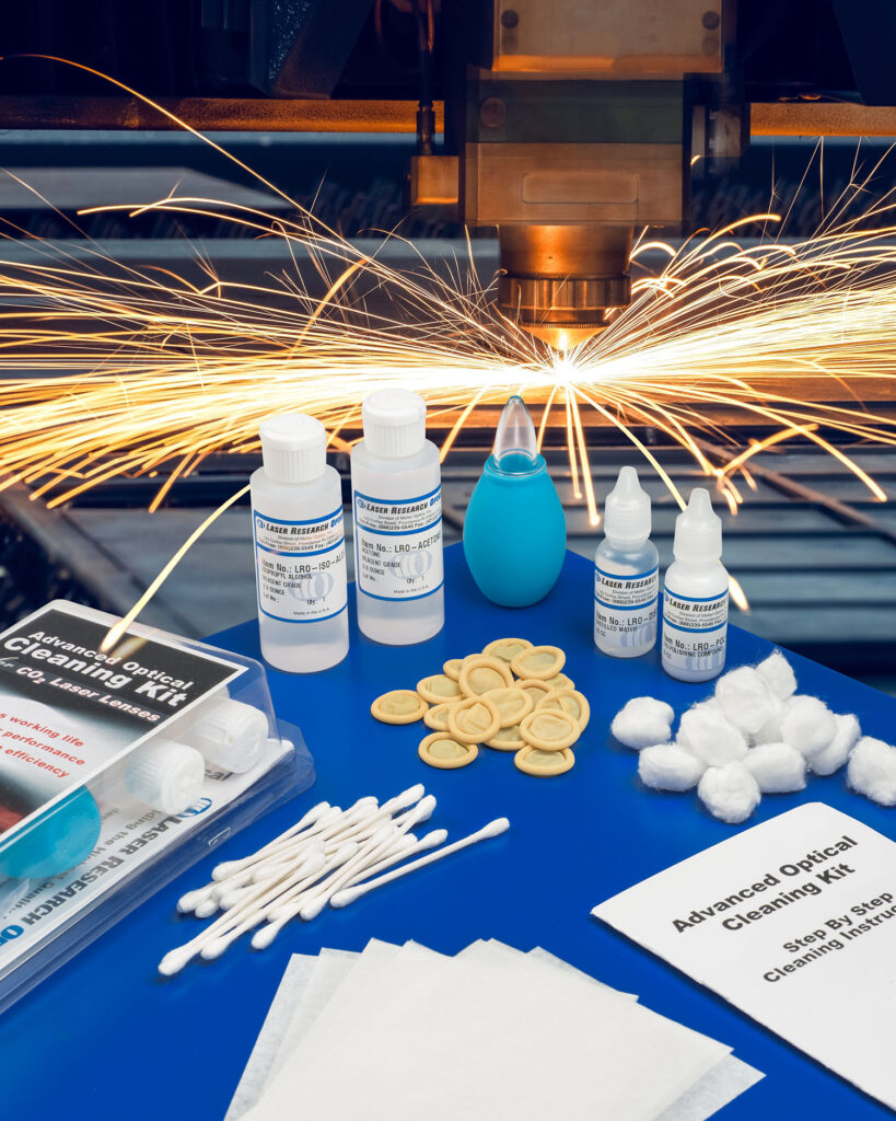 Contents of a laser optic cleaning kit are arranged on a blue table. There are small bottles of isopropyl alcohol, fingercots, cotton swabs and balls and some plastic packaging. A sparking metal cutting laser is in the background.