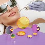 An assortment of Nd:YAG laser optics sit on top a purple table. A woman in the background wearing safety glasses is being treated by a medical laser.