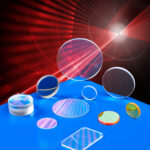 An array of various laser optics in different sizes, shapes, and configurations sit atop a blue table. Red laser lights shine all around them.