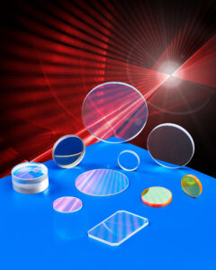 OPTICAL LENSES & WINDOWS FABRICATED TO CUSTOMER SPECIFICATIONS An array of various laser optics in different sizes, shapes, and configurations sit atop a blue table. Red laser lights shine all around them.