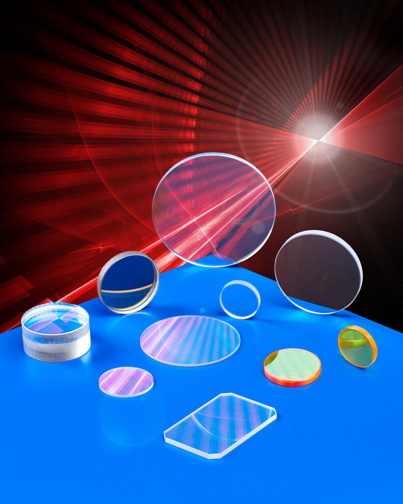 An array of various laser optics in different sizes, shapes, and configurations sit atop a blue table. Red laser lights shine all around them.