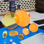 An assortment of laser optics lenses in ZnSe sit on a blue table next to some precision cut metal. A cutting laser is in the background.