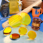 ZnSe lenses for laser engravers sit on a grey surface next to laser mirrors. Engraved awards, plaques and other merchandise are behind them.