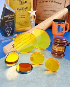 ZnSe lenses for laser engravers sit on a grey surface next to laser mirrors. Engraved awards, plaques and other merchandise are behind them.