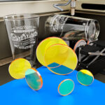 Several Zinc Selenide laser focusing lenses are arranged on a blue table with a laser engraver etching crystal glassware in the background.