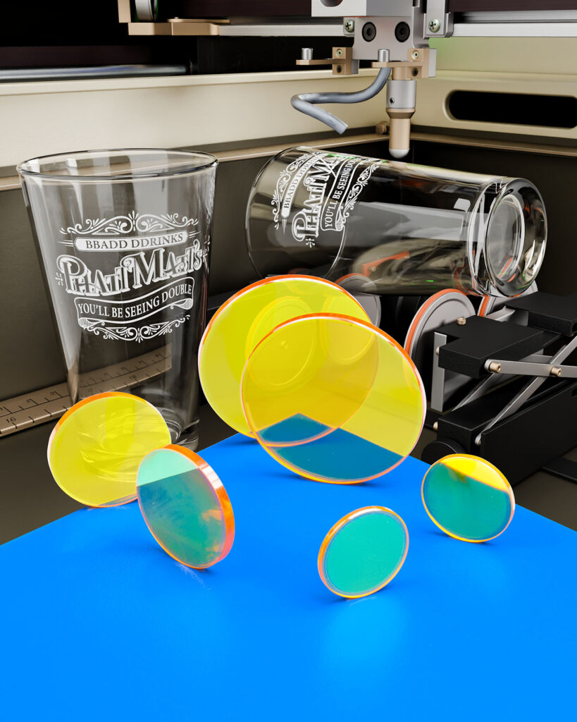 Several Zinc Selenide laser focusing lenses are arranged on a blue table with a laser engraver etching crystal glassware in the background.