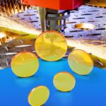 5 ZnSe CO2 laser lenses stand on a blue table. A heavy laser is cutting steel in the background with sparks spraying away from the nozzle.