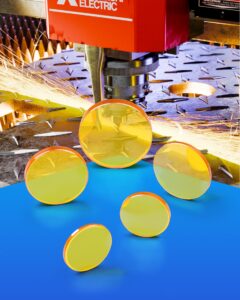 5 ZnSe CO2 laser lenses stand on a blue table. A heavy laser is cutting steel in the background with sparks spraying away from the nozzle.