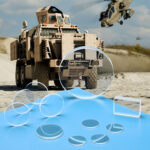 Many Zinc Sulfide windows and lenses in different configurations sit on a blue table in front of military vehicles using guidance systems.