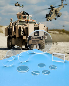 Many Zinc Sulfide windows and lenses in different configurations sit on a blue table in front of military vehicles using guidance systems.
