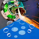 A group of laser debris shields are laying on a blue table in front of an industrial laser emitting sparks.