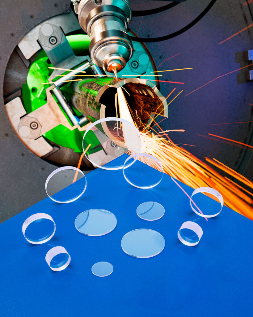 LASER DEBRIS SHIELDS RESIST SPATTER AND PITTING A group of laser debris shields are laying on a blue table in front of an industrial laser emitting sparks.