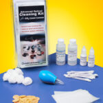 The packaging and contents of a laser optics cleaning kit sit on a blue table in front of a yellow background. Bottles of solution, wipes, cotton swabs, cotton balls and finger cots are spaced evenly.