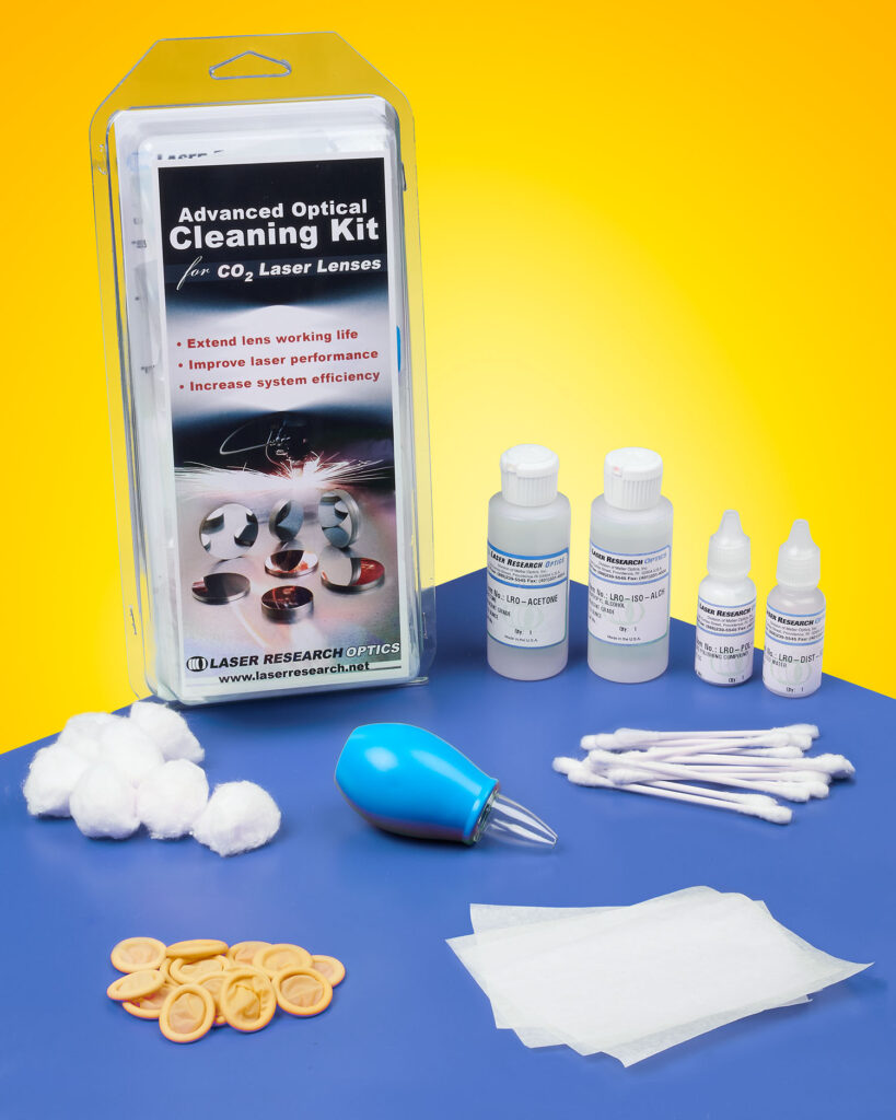 PRECISION OPTICS CLEANING KIT HELPS PRESERVE PERFORMANCE AND LONGEVITY The packaging and contents of a laser optics cleaning kit sit on a blue table in front of a yellow background. Bottles of solution, wipes, cotton swabs, cotton balls and finger cots are spaced evenly.