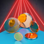 A handful of CO2 laser beam delivery optics sit on a blue table with an array of red lasers in the background. Reminiscent of a very cool style of 1980s yearbook photo.