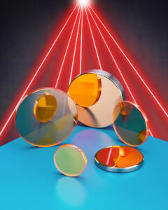 A handful of CO2 laser beam delivery optics sit on a blue table with an array of red lasers in the background. Reminiscent of a very cool style of 1980s yearbook photo.