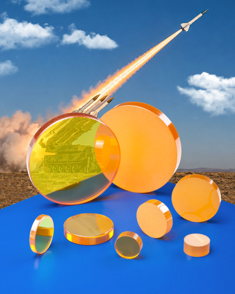 Several zinc selenide laser optics sit in front of a photo with a rocket launching.